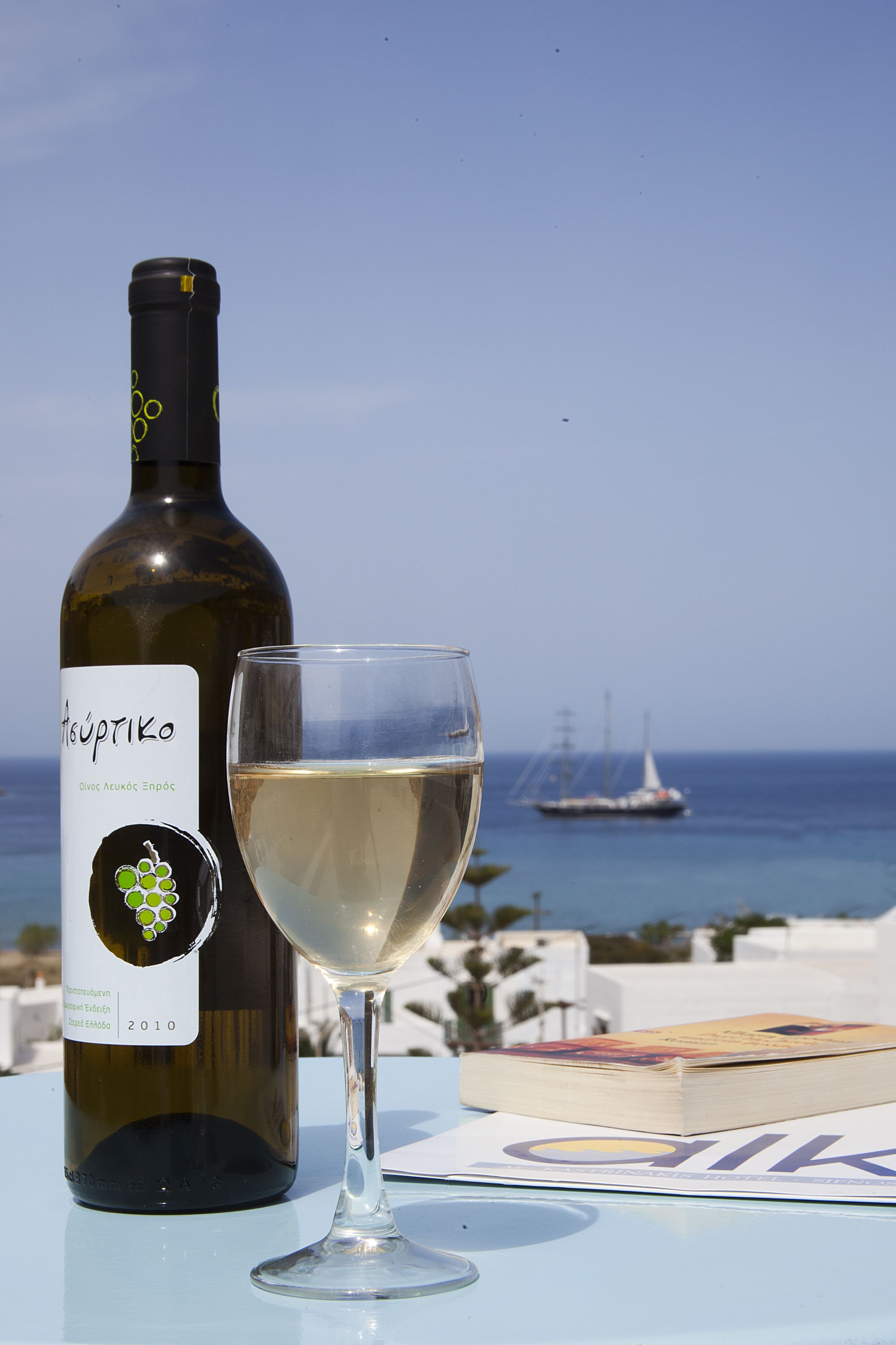 En flaska Oivoc Acouas Enpoc-vin och ett glas vitt vin står på ett bord tillsammans med en bok. I bakgrunden syns havet och några båtar.