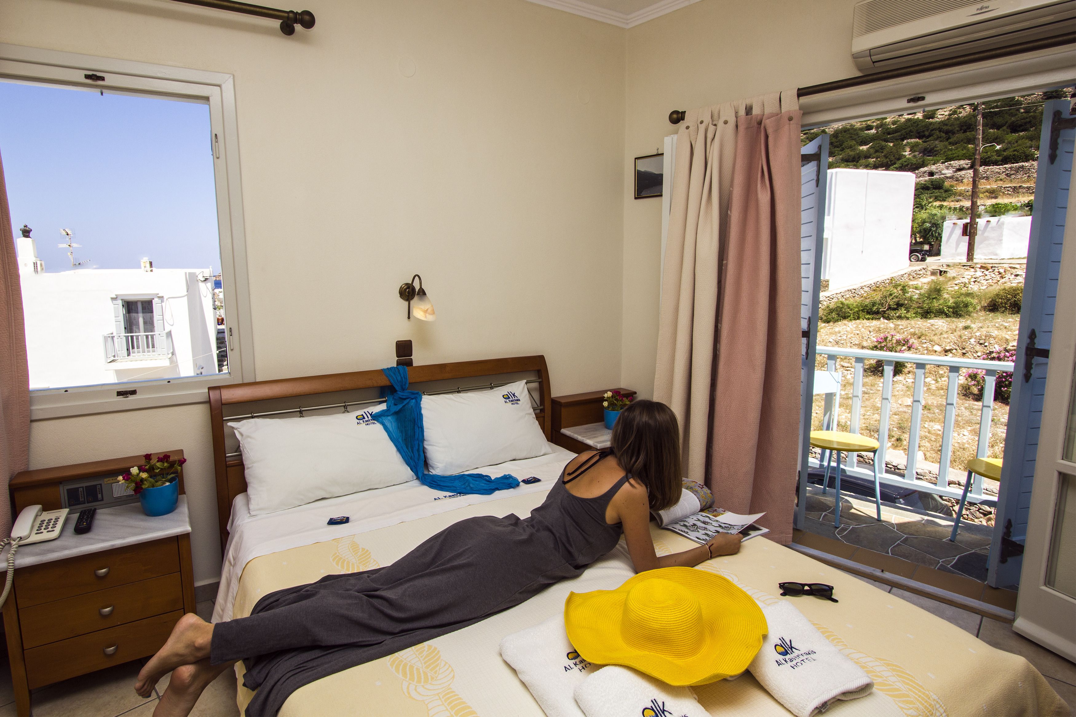 Ein Hotelzimmer mit Bett, Nachttisch mit Telefon, Fenster mit Blick auf Gebäude und Balkon mit Geländer.