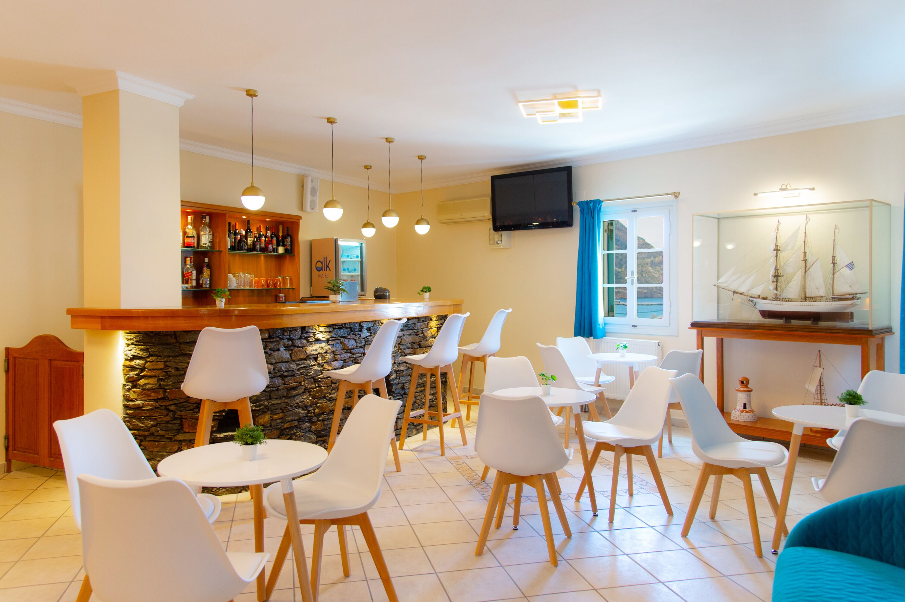 Zona de bar con pared de piedra, sillas blancas y mostrador de madera. Hay un televisor montado en la pared y un modelo de barco sobre una mesa.