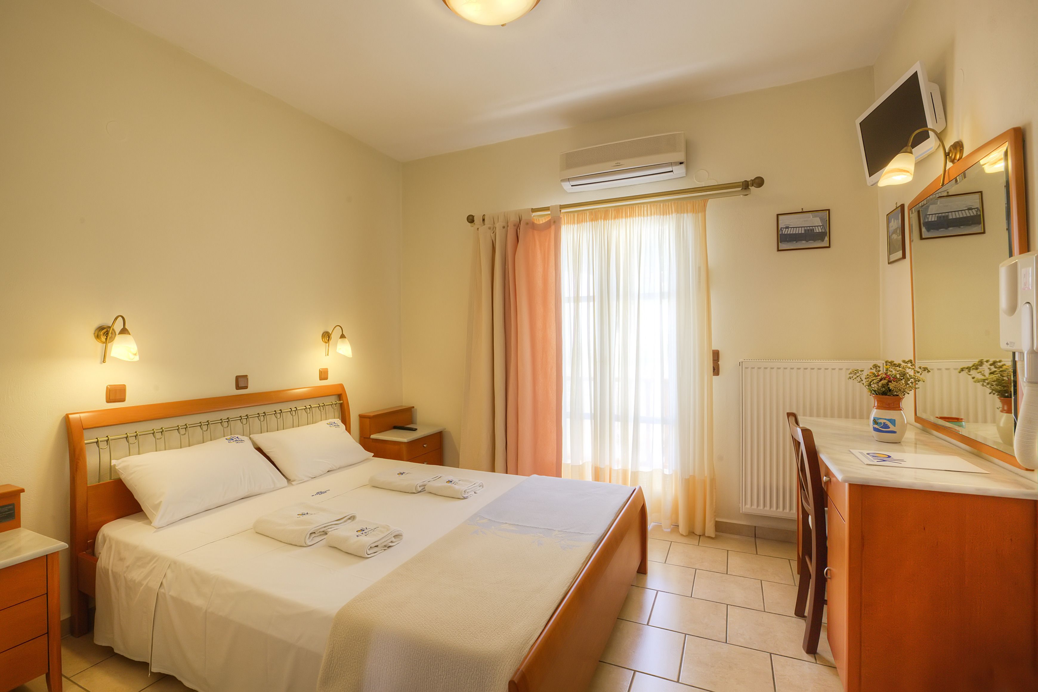 A hotel room with a bed, bedside tables, a desk, a mirror, and an air conditioning unit.