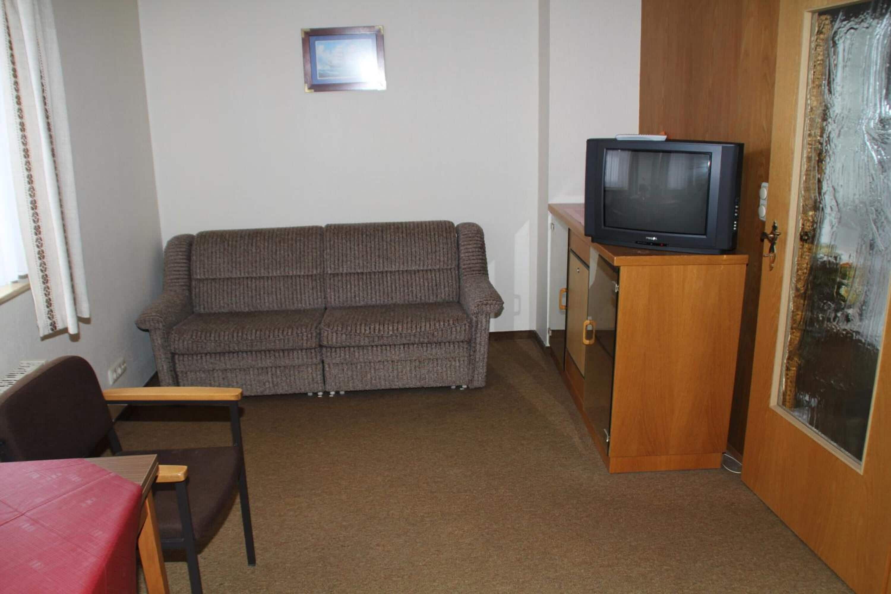 Uma sala de estar com um sofá marrom, um móvel de televisão de madeira, uma televisão, uma mesa vermelha e uma cadeira.