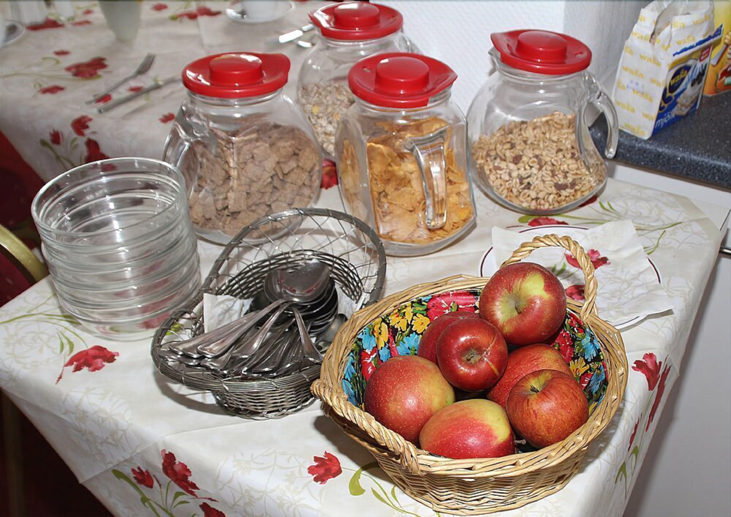 A table with jars of granola, a basket of apples, and a stack of clear glasses.
