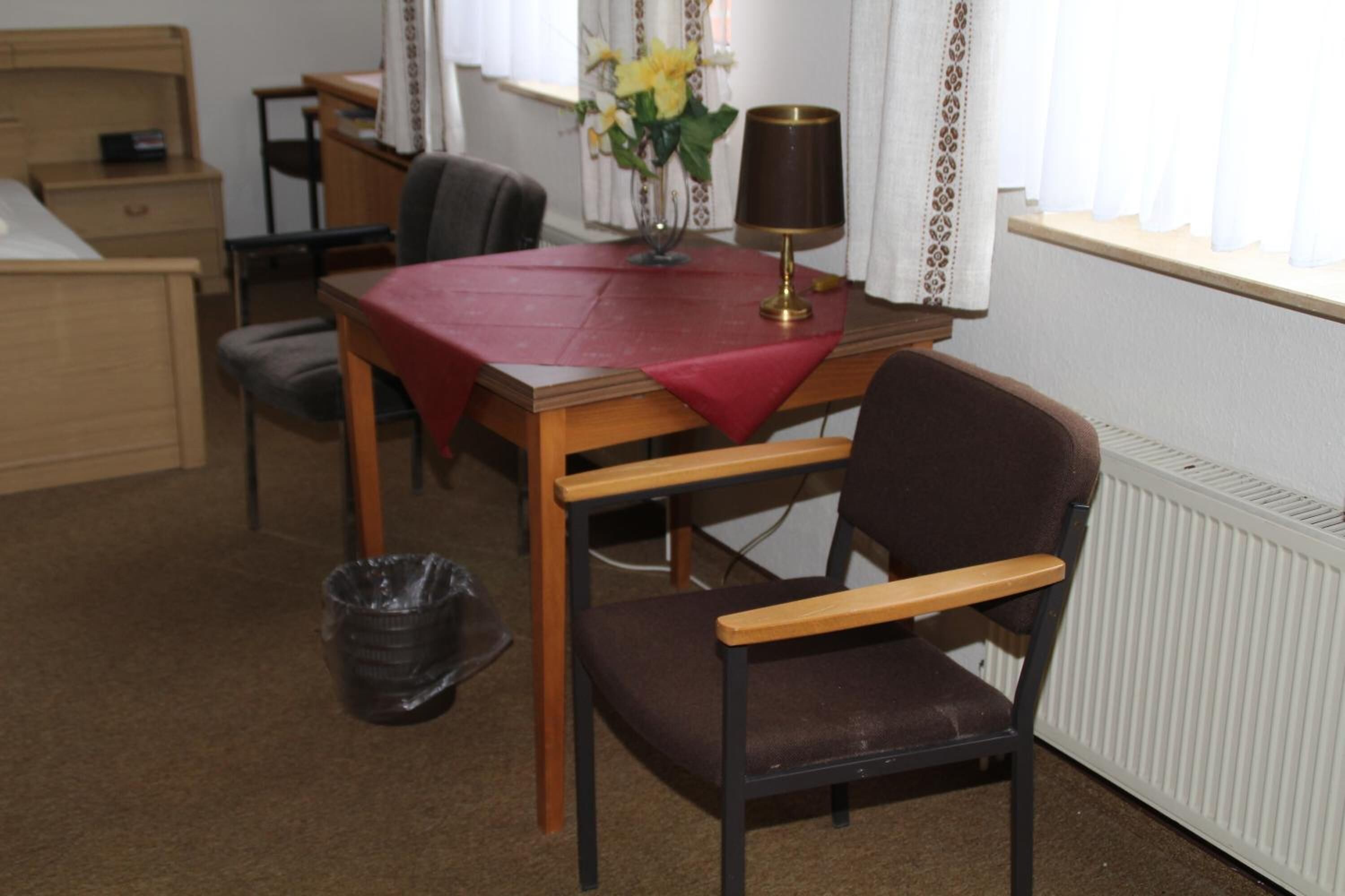 Et lille, møbleret rum med et træskrivebord, en stol, et bord med rødt dug, en lampe og en radiator.