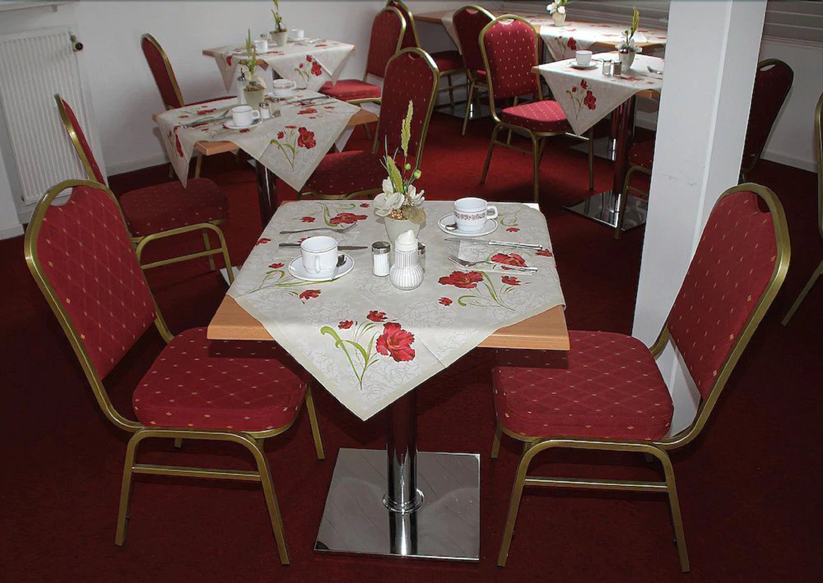 Ein Essbereich mit Tischen für zwei Personen, jeweils mit roten Sitzmöbeln und weißen Tischtüchern mit Blumenmuster.