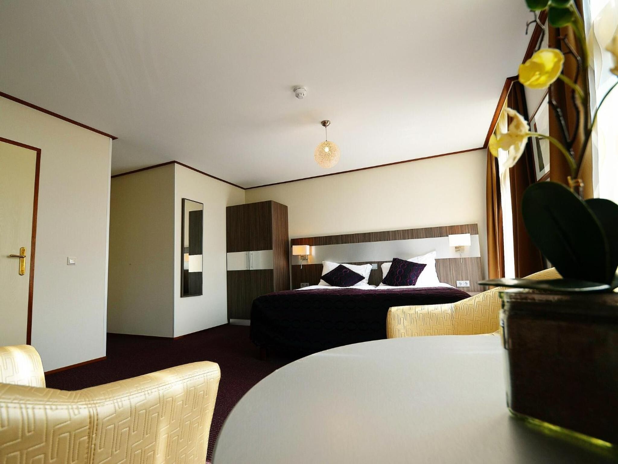 Una habitación de hotel con cama, mesita de noche, silla, espejo y lámpara colgante.
