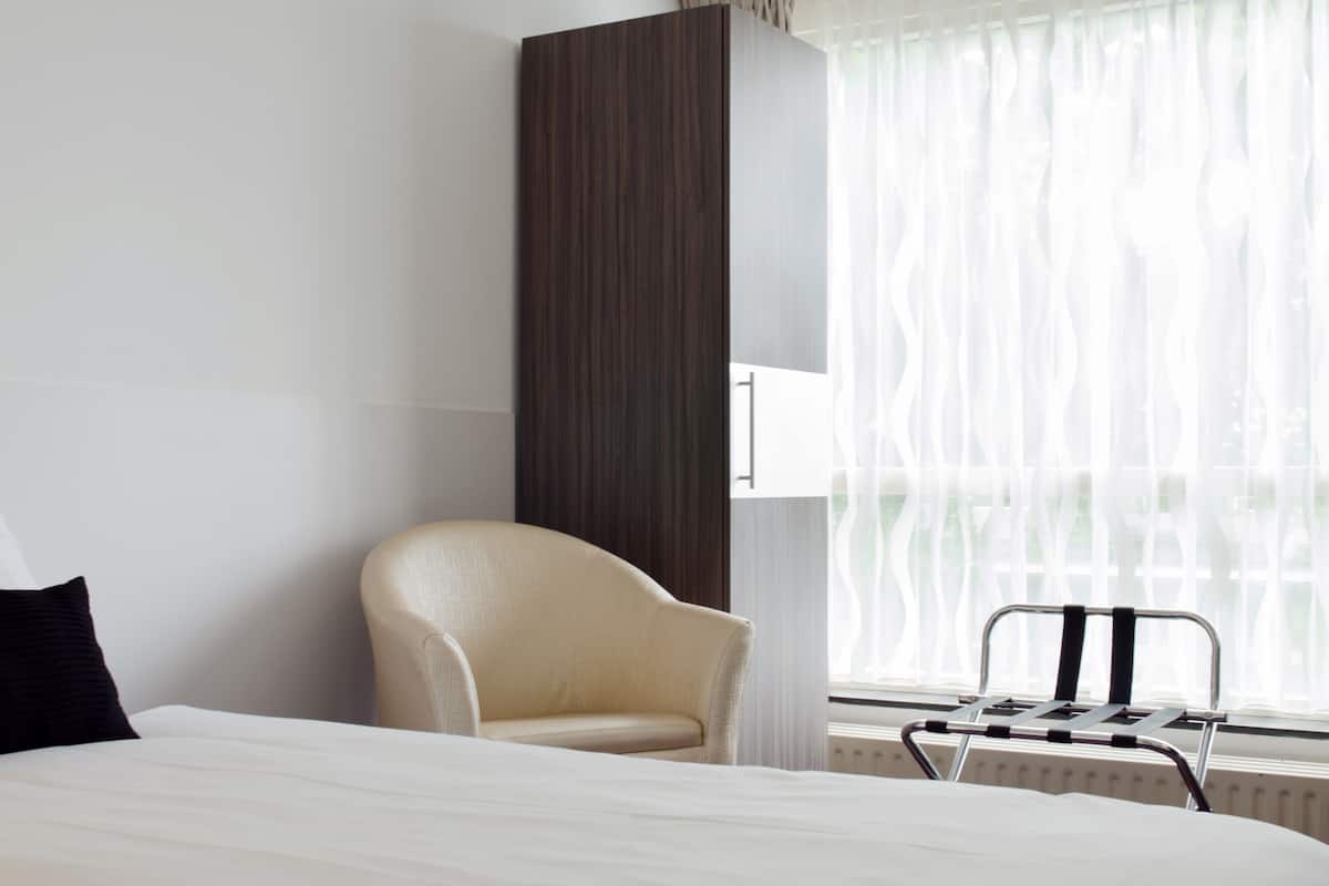 A hotel room with a bed, a chair, a radiator, and a window with sheer curtains.