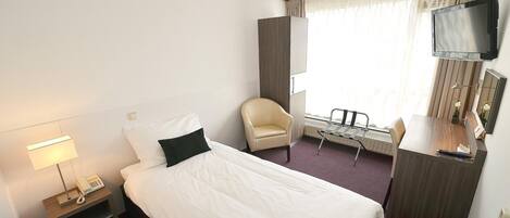 Ein Hotelzimmer mit einem Einzelbett, einem Schreibtisch, einem Stuhl, einem Fernseher und einem Fenster mit Vorhängen.