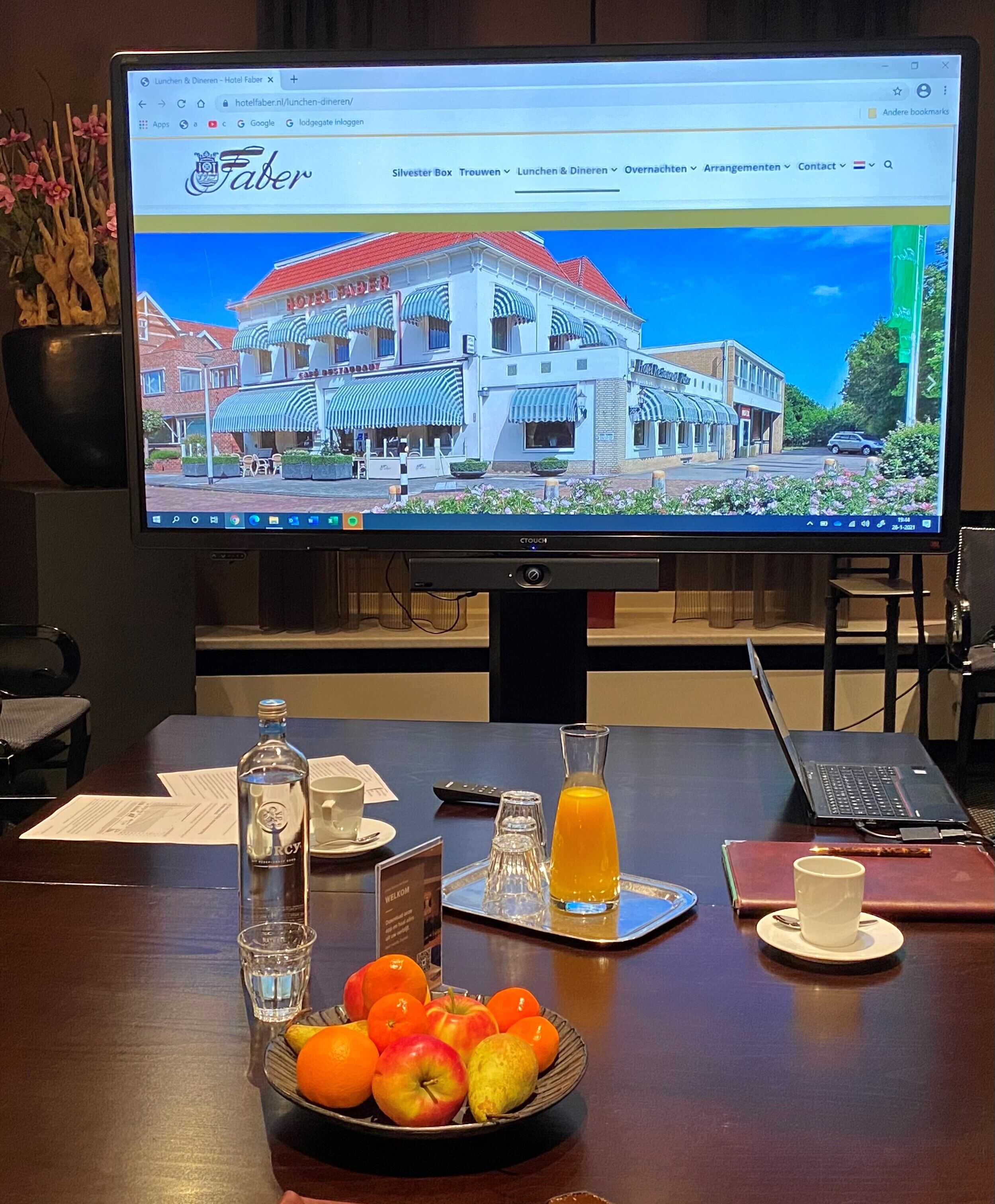 Ein Hotelzimmer mit einem großen Bildschirm, der die Hotelwebsite zeigt, einem Holztisch mit einer Obstschale, einem Glas Orangensaft und einem Laptop.