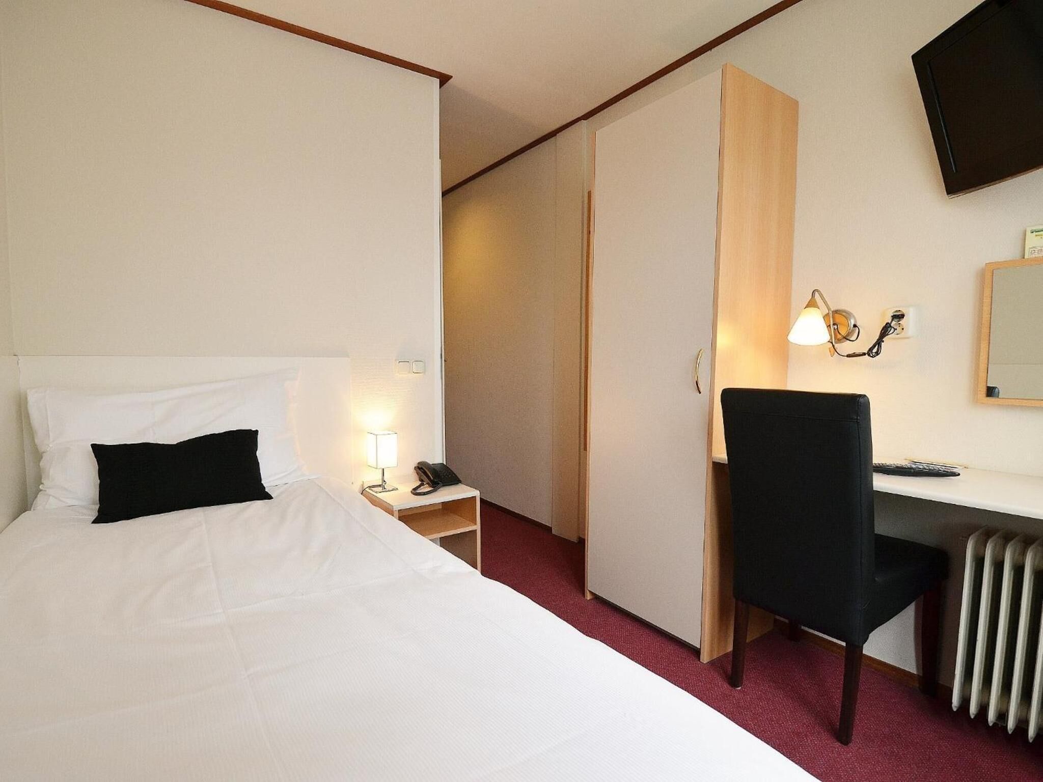 A hotel room with a bed, a desk, a chair, a lamp, a phone, a TV, and a radiator.
