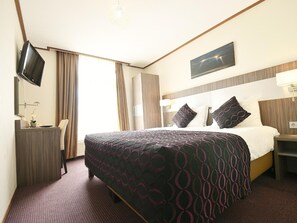 A hotel room with a large bed, a flat-screen TV mounted on the wall, a desk with a lamp, and a window with curtains. - Hotel Faber (Hoogezand)
