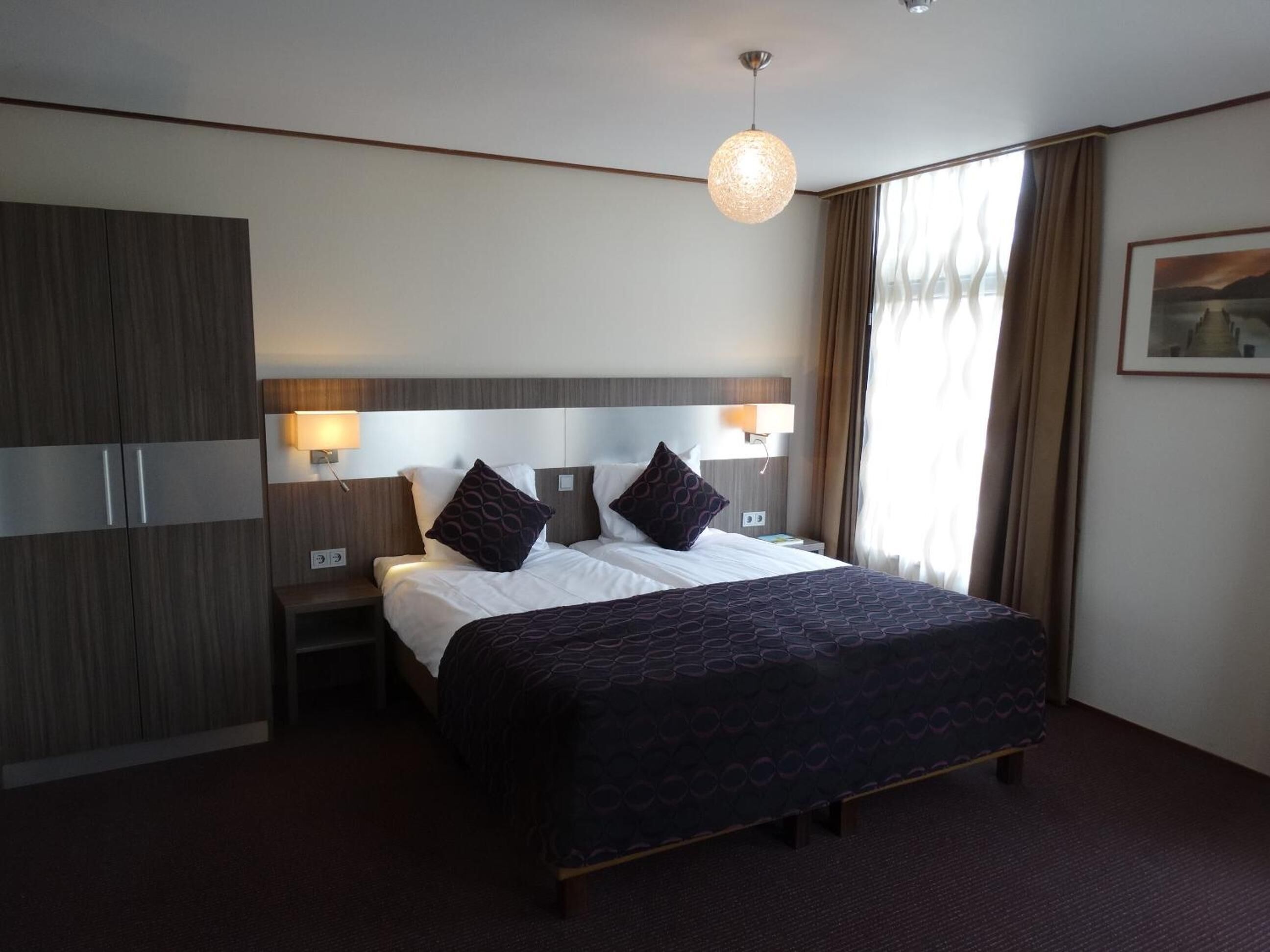 Una habitación de hotel con una cama grande, dos mesitas de noche, un armario y una lámpara colgante.