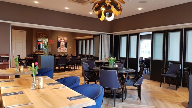 A spacious dining area with multiple tables and chairs, a wooden floor, and a modern chandelier. - Hotel Faber (Hoogezand)