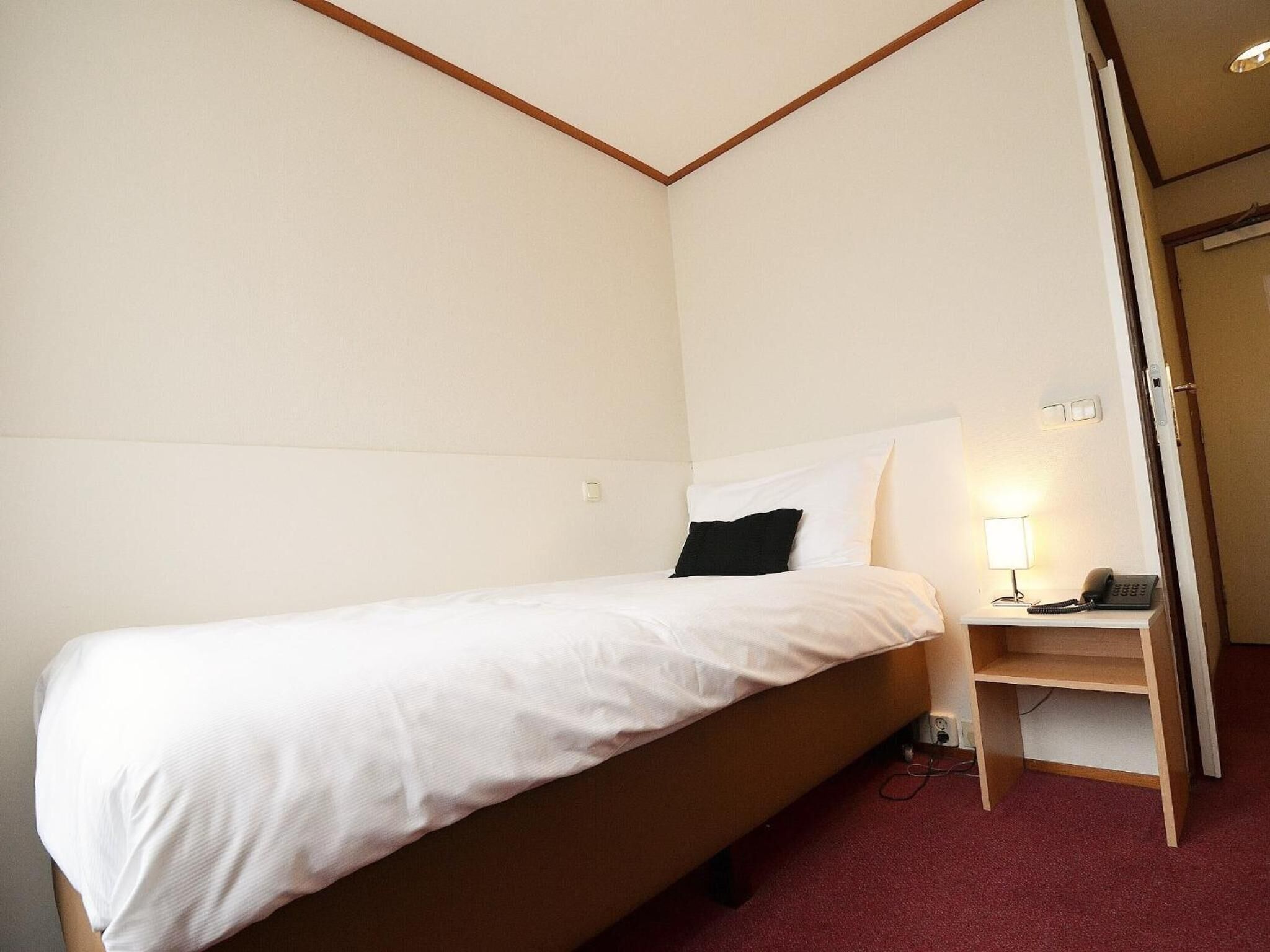 A single bed with white bedding in a room with a wooden headboard and a small bedside table with a lamp and phone.