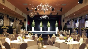 A banquet hall with round tables, white chairs, and a stage with a backdrop. - Hotel Faber (Hoogezand)