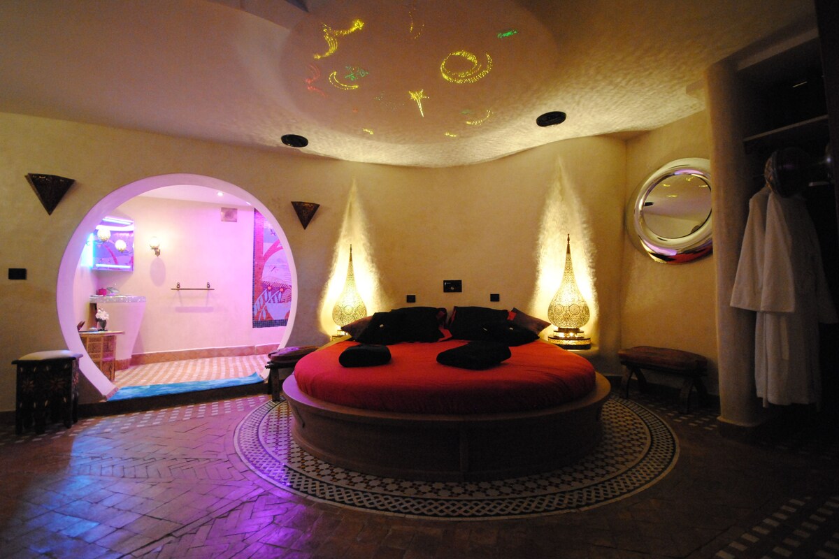 Un lit rond avec un matelas rouge, dans une pièce dont le plafond présente un design unique et qui est agrémentée de deux lampes fixées au mur.