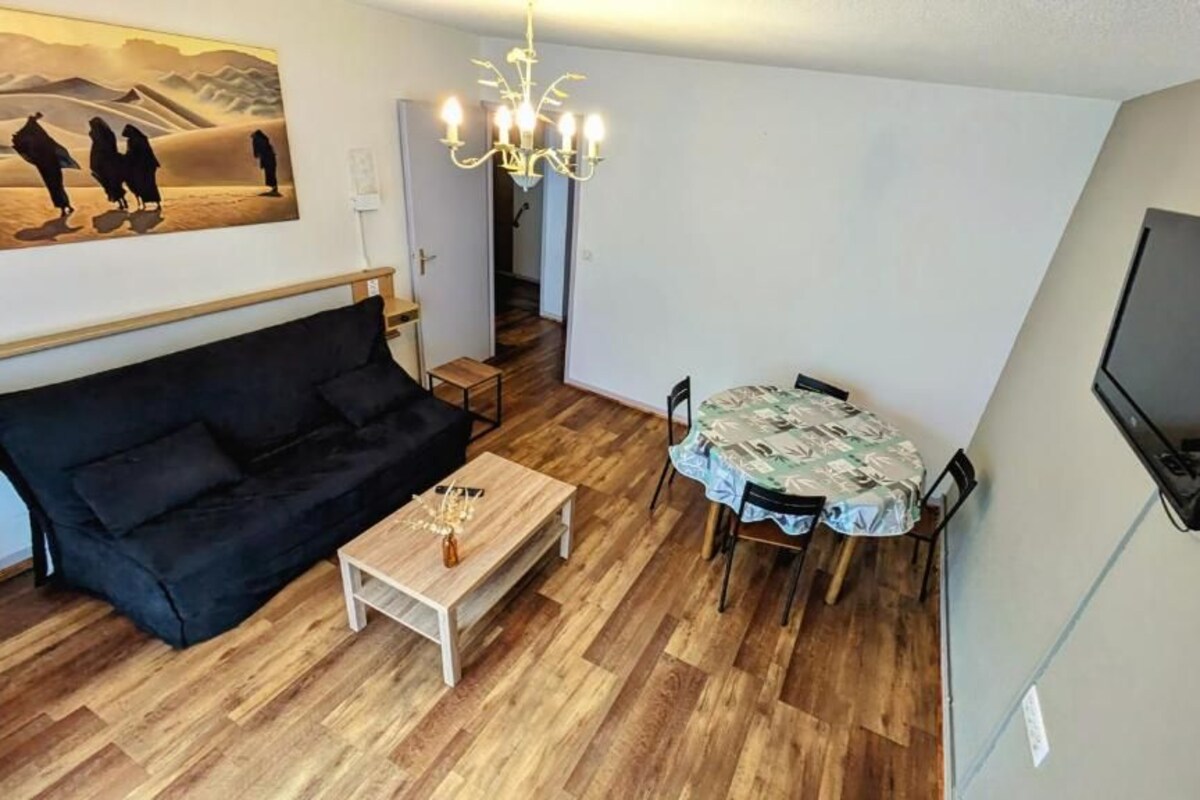 Un salon avec un canapé noir, une table basse en bois, une table à manger avec des chaises, un lustre et un téléviseur à écran plat fixé au mur.