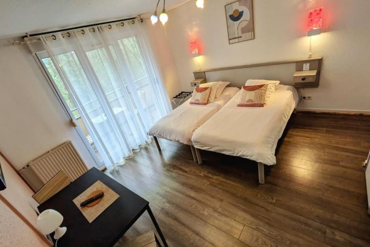 Une chambre d’hôtel avec deux lits, une table avec une chaise, un radiateur et une fenêtre avec des rideaux.