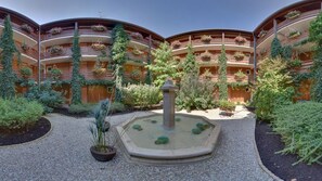 Une cour intérieure avec une fontaine centrale, entourée d’une végétation luxuriante et d’un bâtiment doté de nombreux balcons ornés de fleurs.