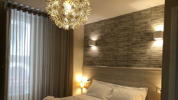 A hotel room with a bed, a chandelier, a headboard with a textured wall, and a window with curtains. - Elios Rooms (Catania)