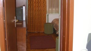 Een smalle gang die leidt naar een slaapkamer met een bed, nachtkastje, radiator en een raam met gordijnen.