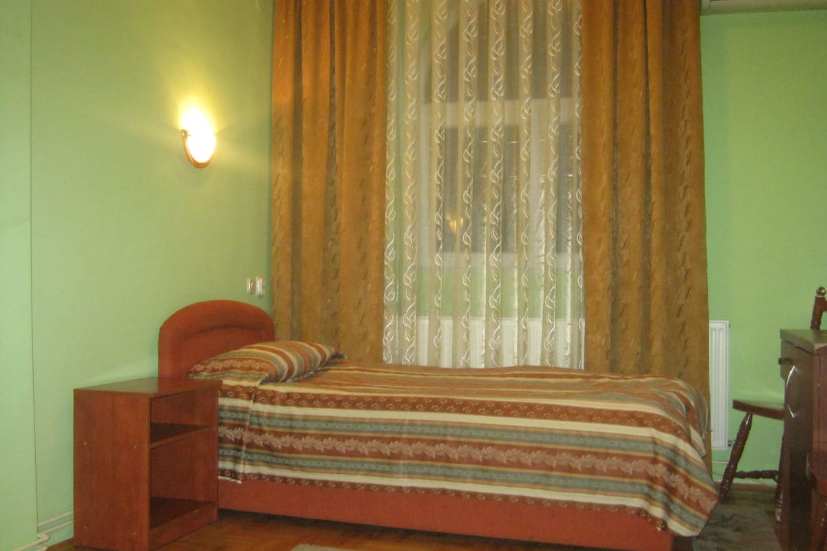 A bedroom with a bed, a nightstand, a chair, a radiator, and a window with curtains.