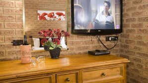 A room with a brick wall, a wooden dresser, a flat-screen TV mounted on the wall, a potted plant, and a pink bottle. - Beechworth Carriage Motor Inn (Beechworth)
