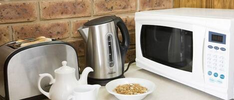 A kitchen with a toaster, kettle, microwave, teapot, and a bowl of cereal.