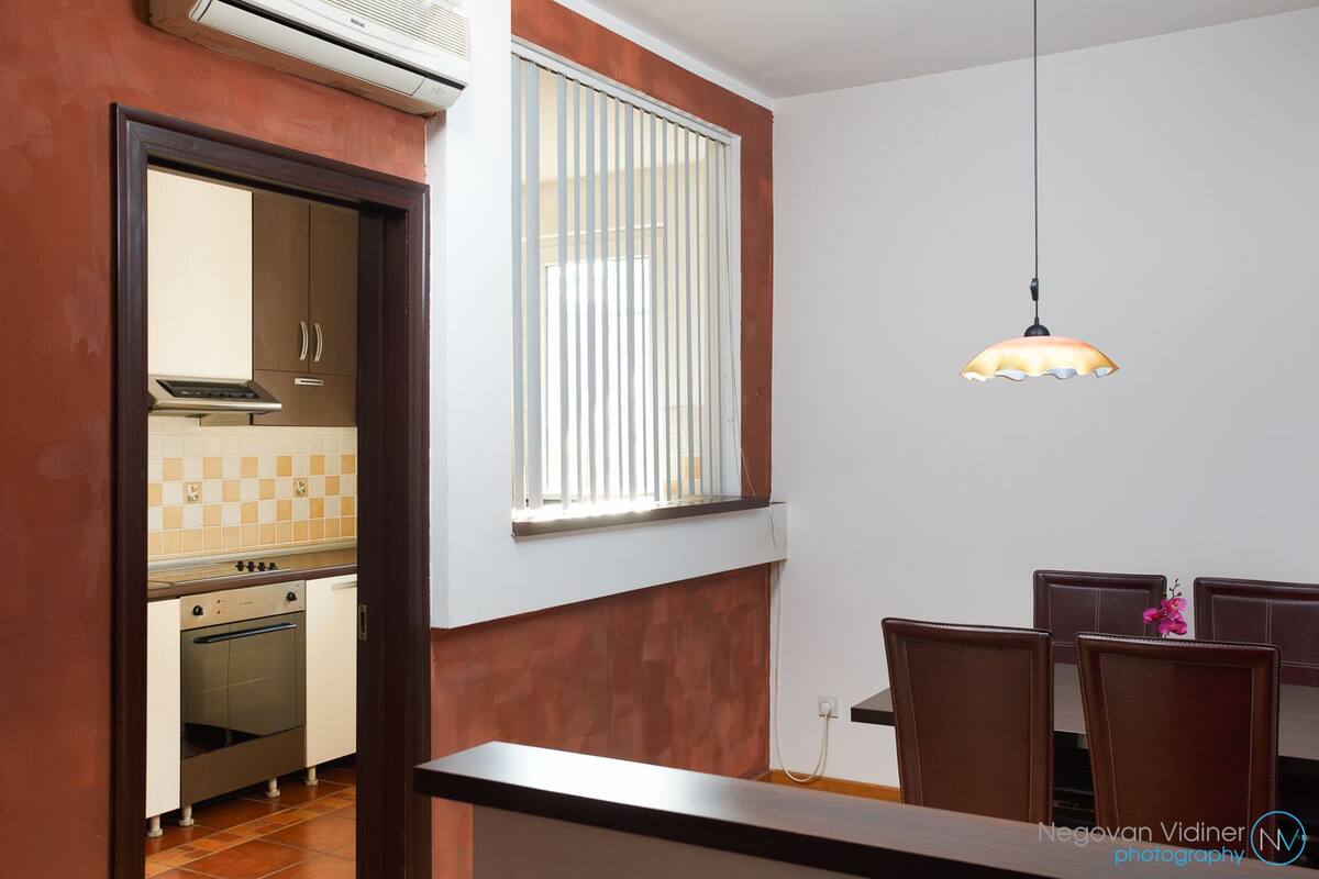 A kitchen with a stove, oven, and window with blinds. There is a dining area with chairs and a pendant light.
