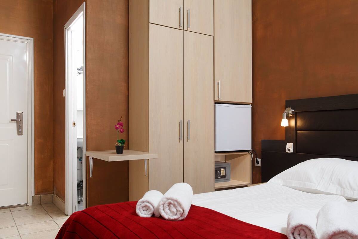 A hotel room with a bed, a nightstand, a TV, a wardrobe, and a small shelf with a plant.