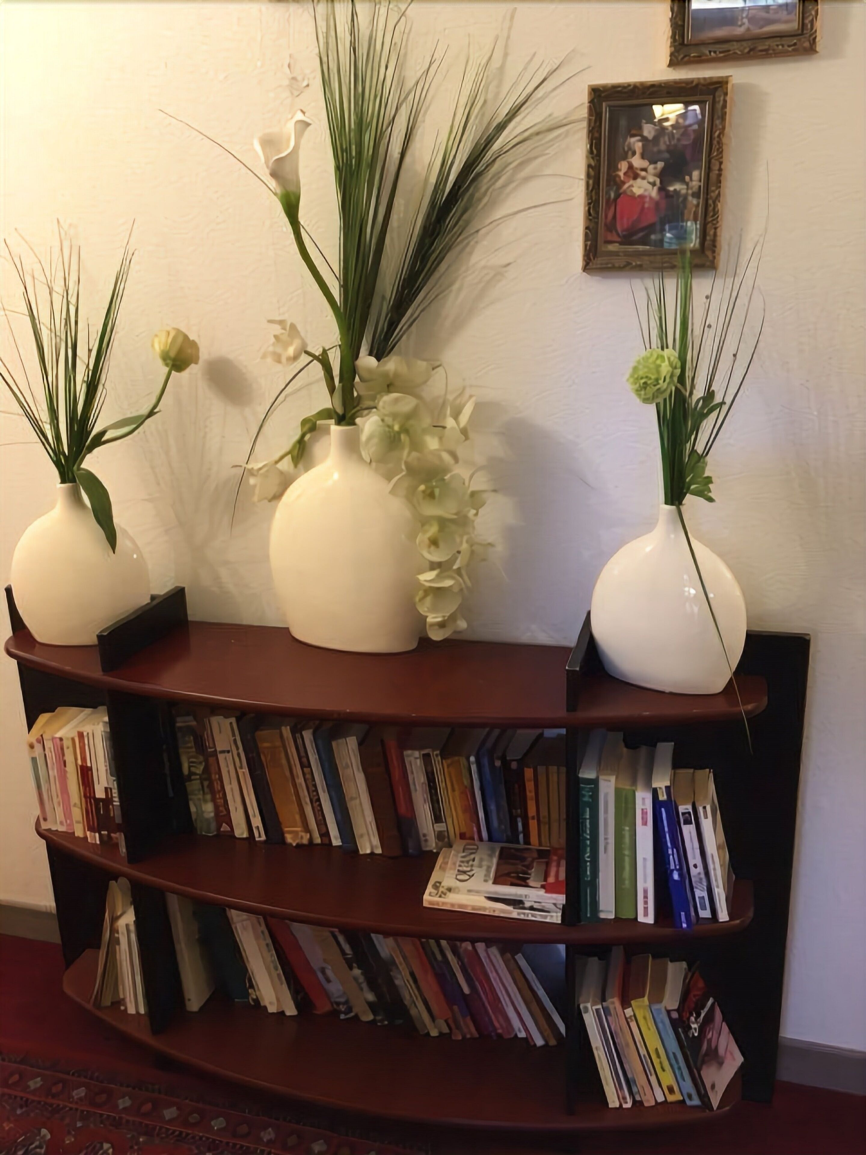 A bookshelf with books and three white vases with flowers.