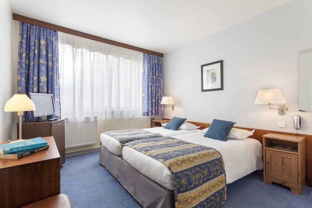 Habitación de hotel con dos camas, un escritorio, una silla, una lámpara, un cuadro en la pared y una ventana con cortinas.
