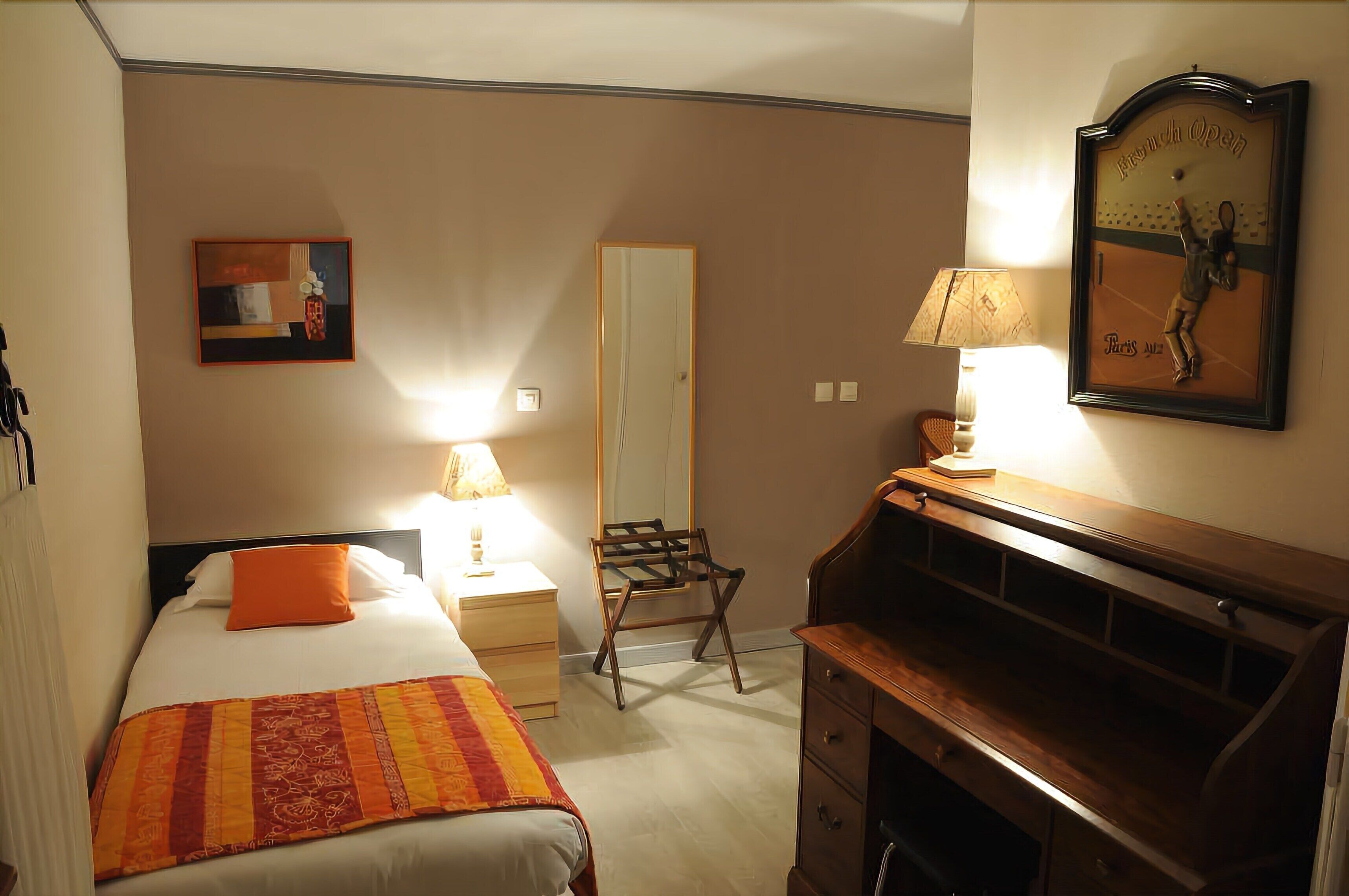 A hotel room with a bed, a desk, a chair, a lamp, a mirror, and a painting on the wall.