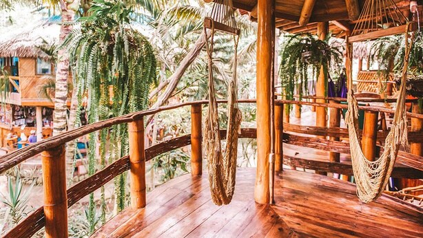 Una terraza de madera con techo de paja, barandillas de bambú y hamacas colgantes.