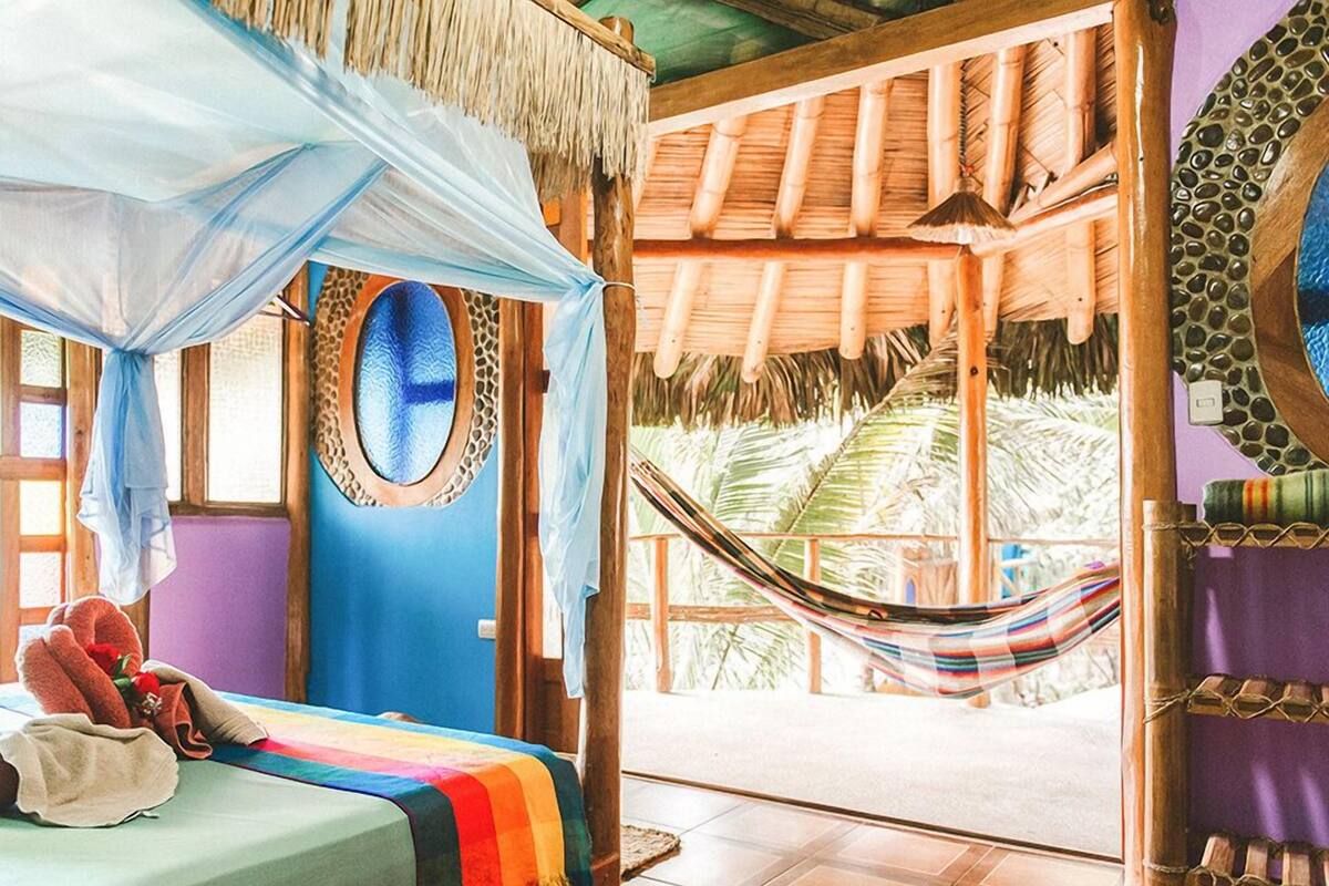 A room with a bed, a hammock, a window with a view of palm trees, and a thatched roof.