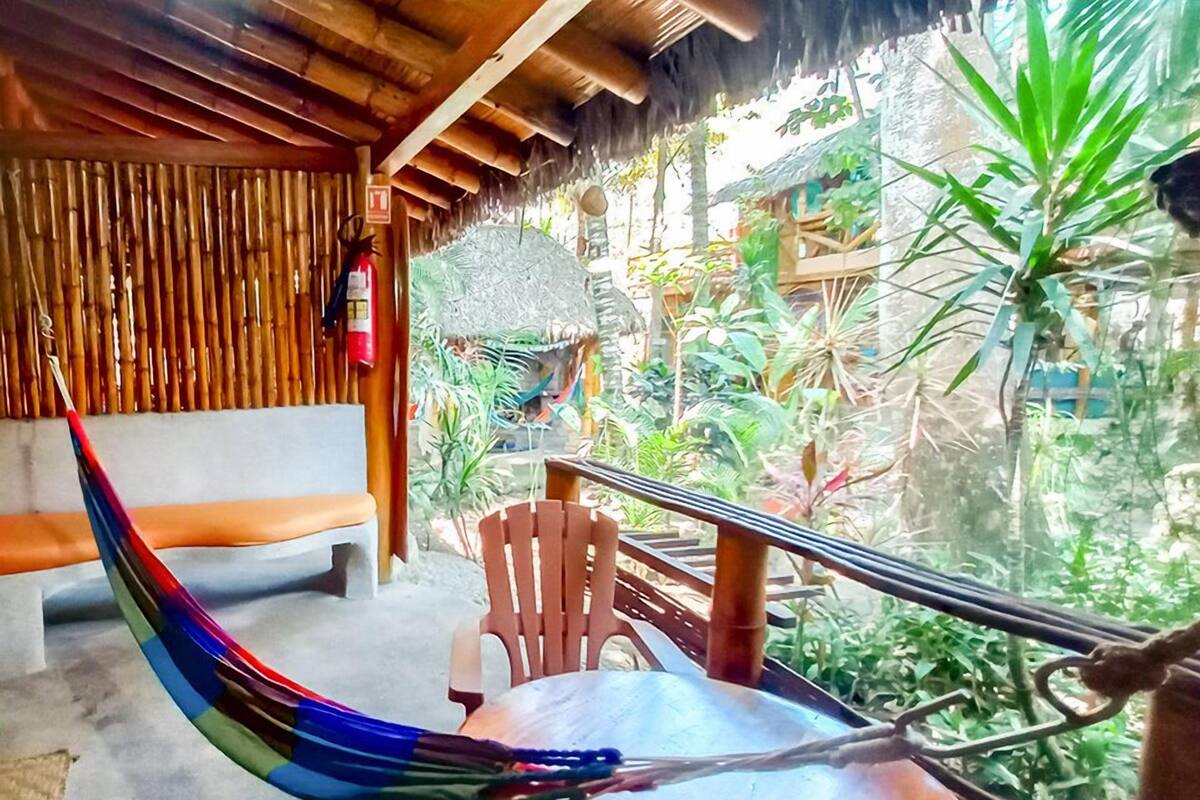 A hammock chair, wooden bench, and a table in a covered outdoor area with a view of lush greenery.
