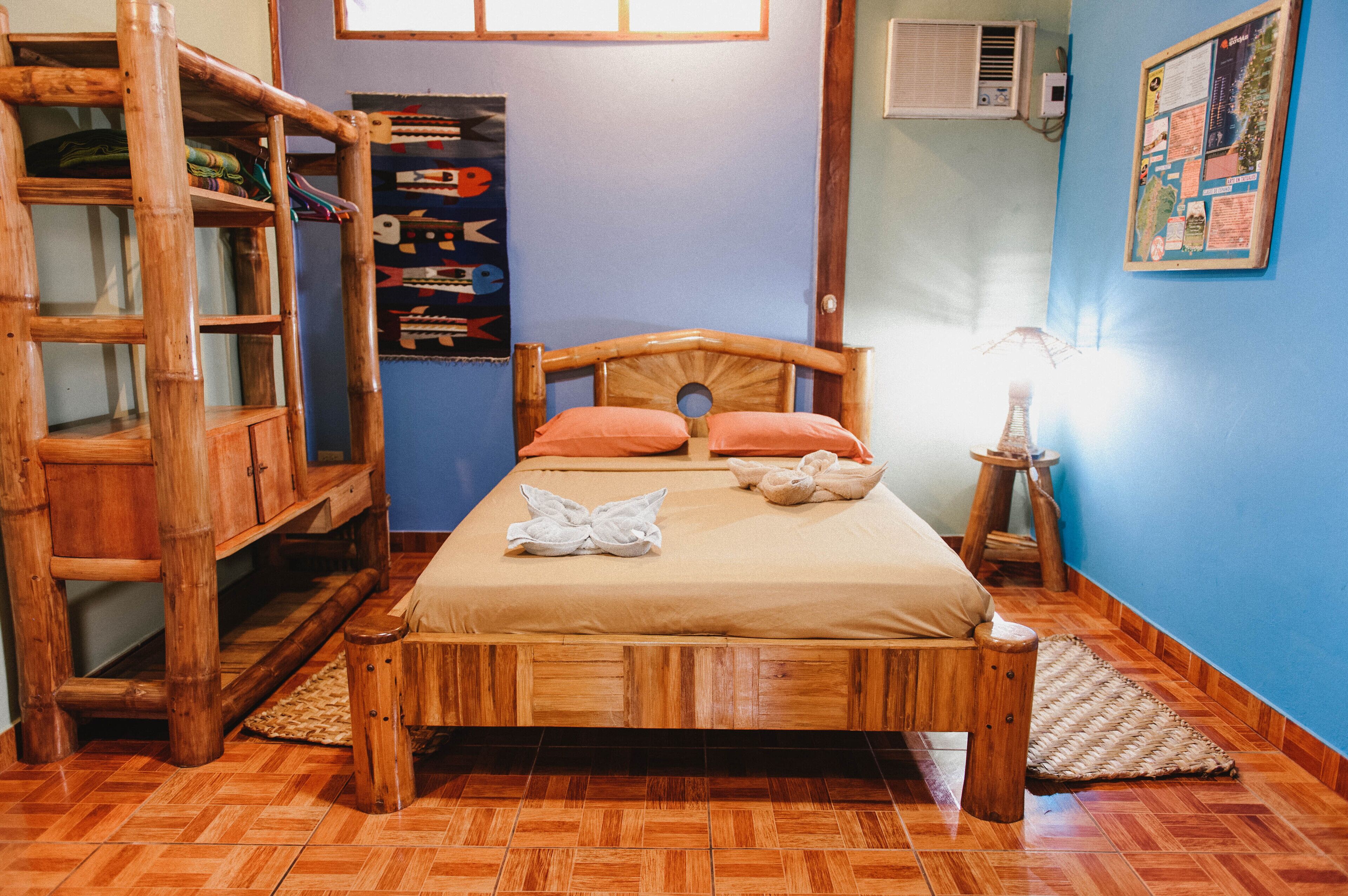 Un lit en bois avec une tête de lit, deux oreillers et une couverture. Il y a une armoire en bois à gauche et une petite table avec une lampe à droite.