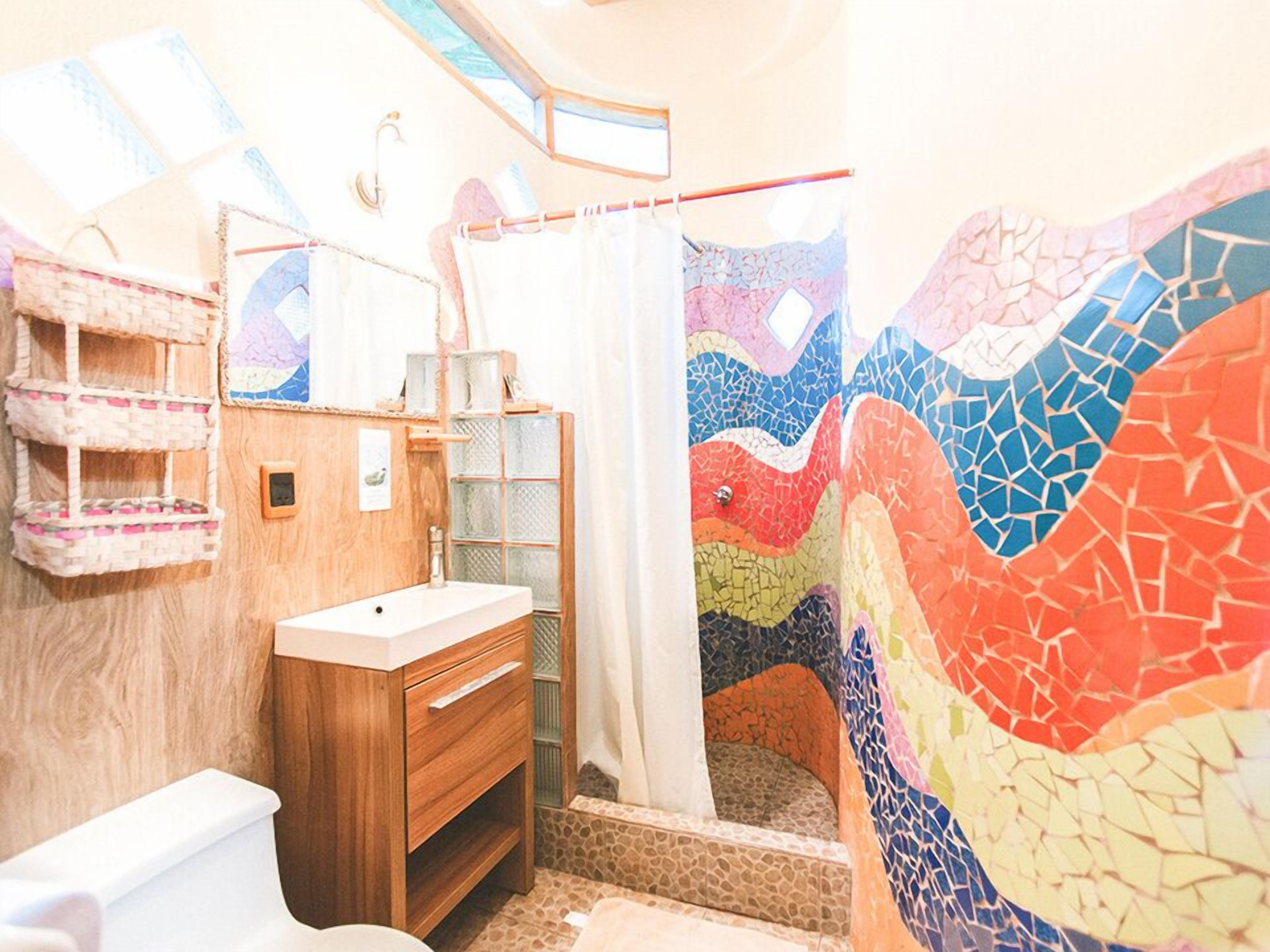 A bathroom with a wooden vanity, a white sink, a shower with a colorful mosaic wall, and a small shelf with towels.