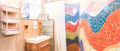 A bathroom with a wooden vanity, a white sink, a shower with a colorful mosaic wall, and a small shelf with towels.