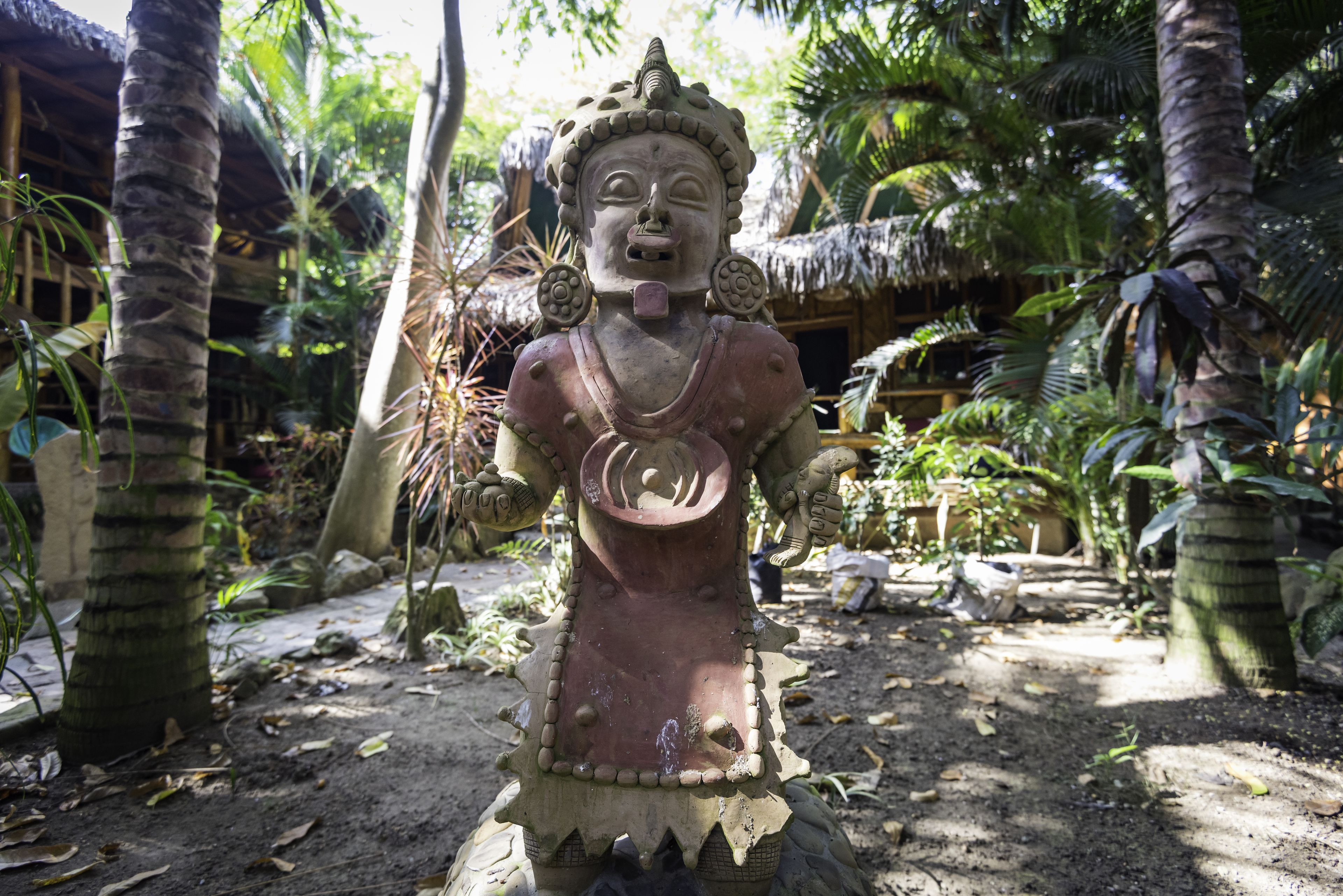 Una estatua en un jardín tropical con palmeras y una estructura de techo de paja al fondo.