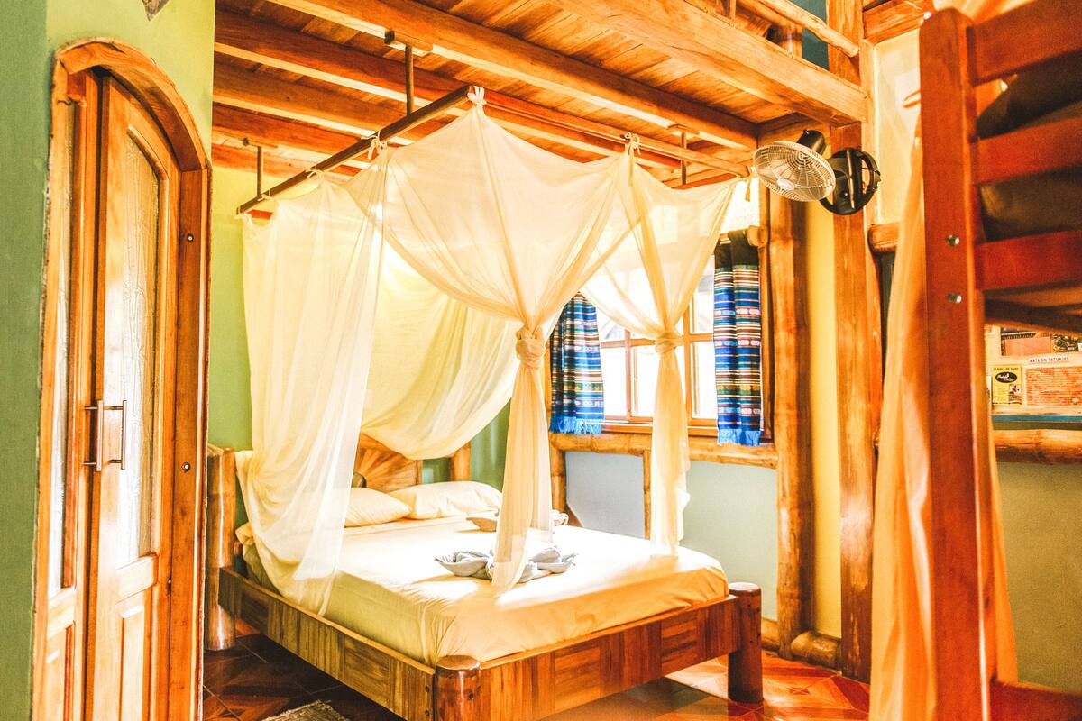 A wooden four-poster bed with a canopy, a wooden door, and a window with striped curtains.