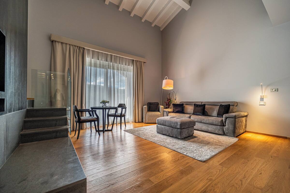 A modern living room with a grey sofa, a small ottoman, a dining table with chairs, and a staircase leading to an upper floor.