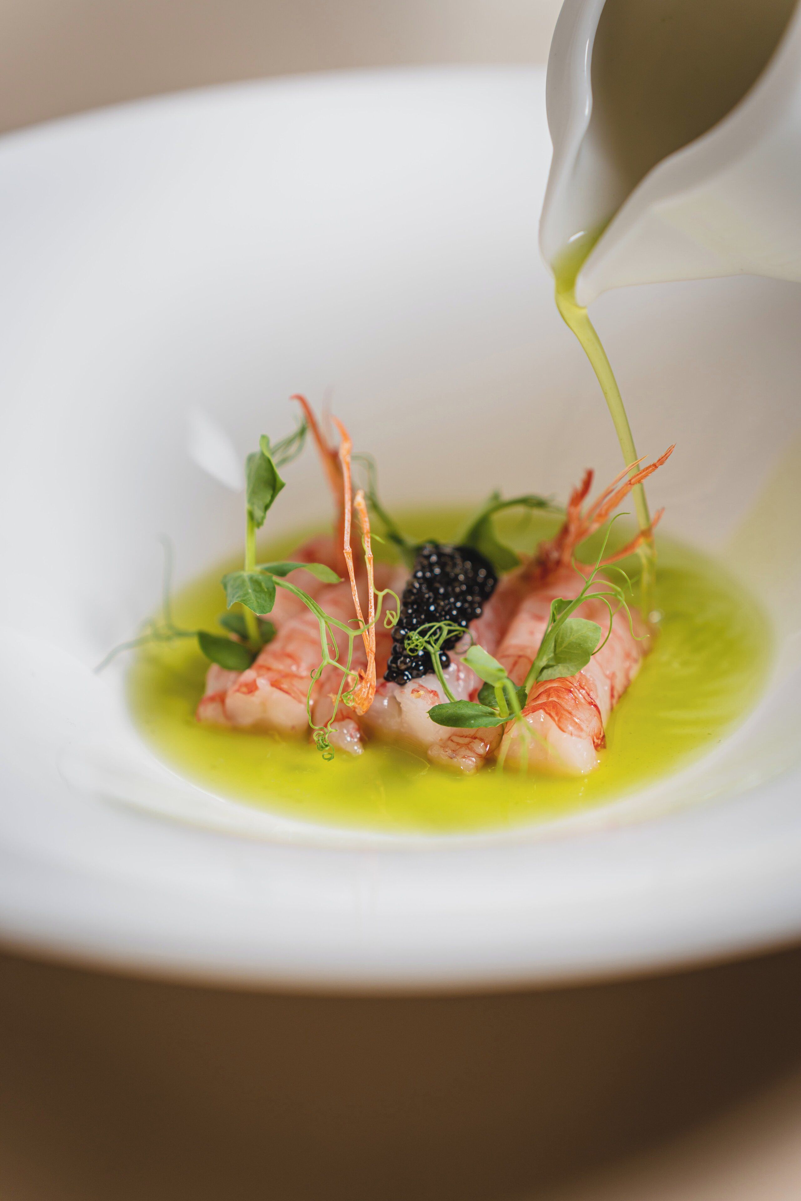 A white bowl with a green sauce being poured over a dish containing fish and garnishes.