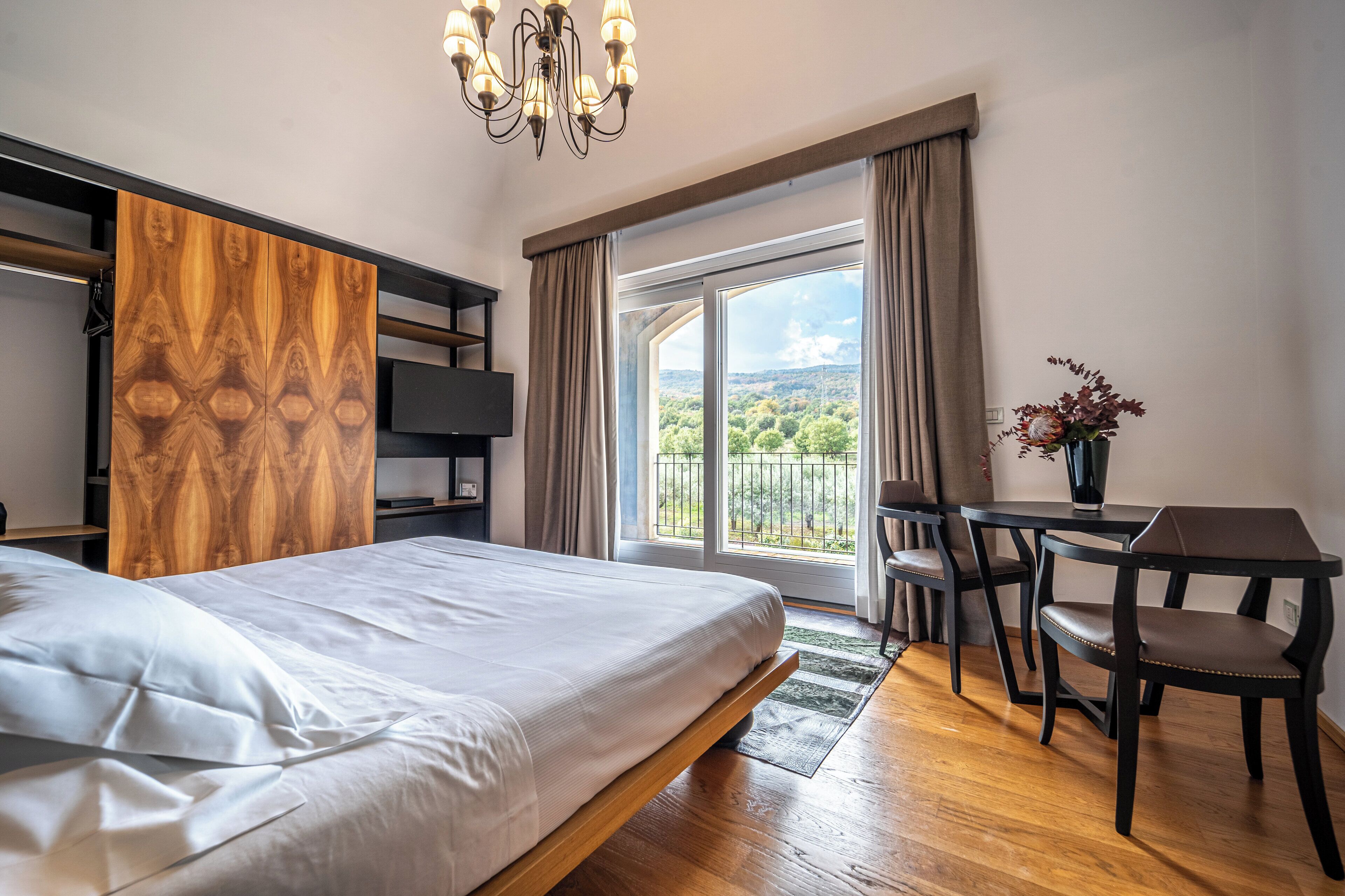 A bedroom with a large bed, a wooden headboard, a desk with a chair, and a view of greenery through the window.