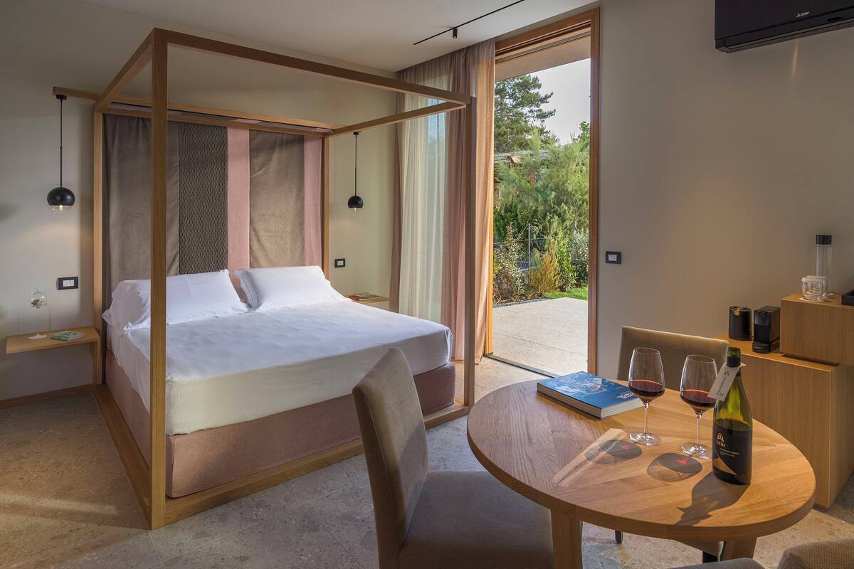 A modern hotel room with a bed, a dining table with wine glasses, and a view of greenery outside.