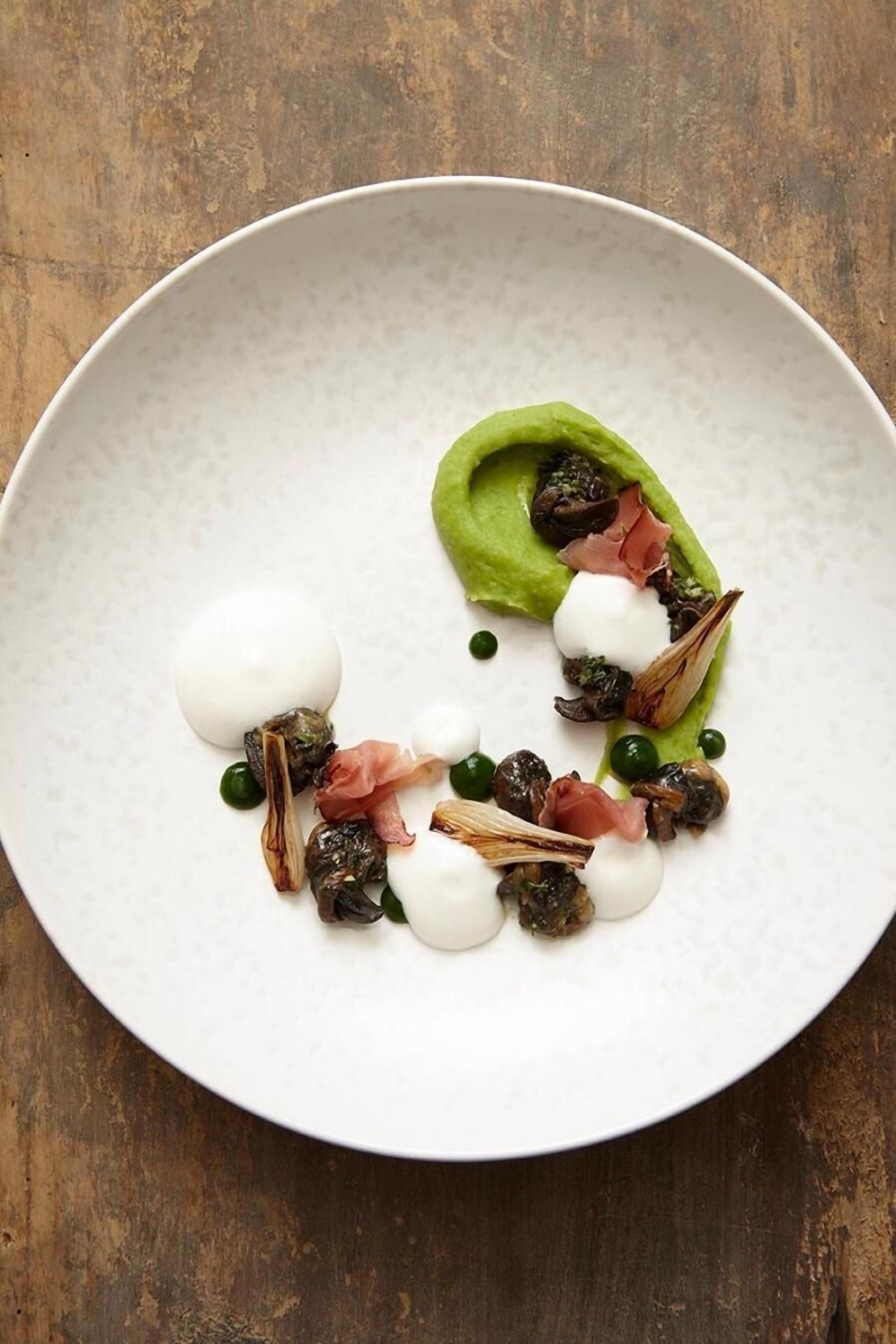 Un plato blanco con salsa verde, esferas blancas, guisantes verdes y hierbas secas.