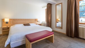 Hotelkamer met een groot bed, houten hoofdeinde, een bankje, nachtkastje, spiegel en raam met gordijnen.