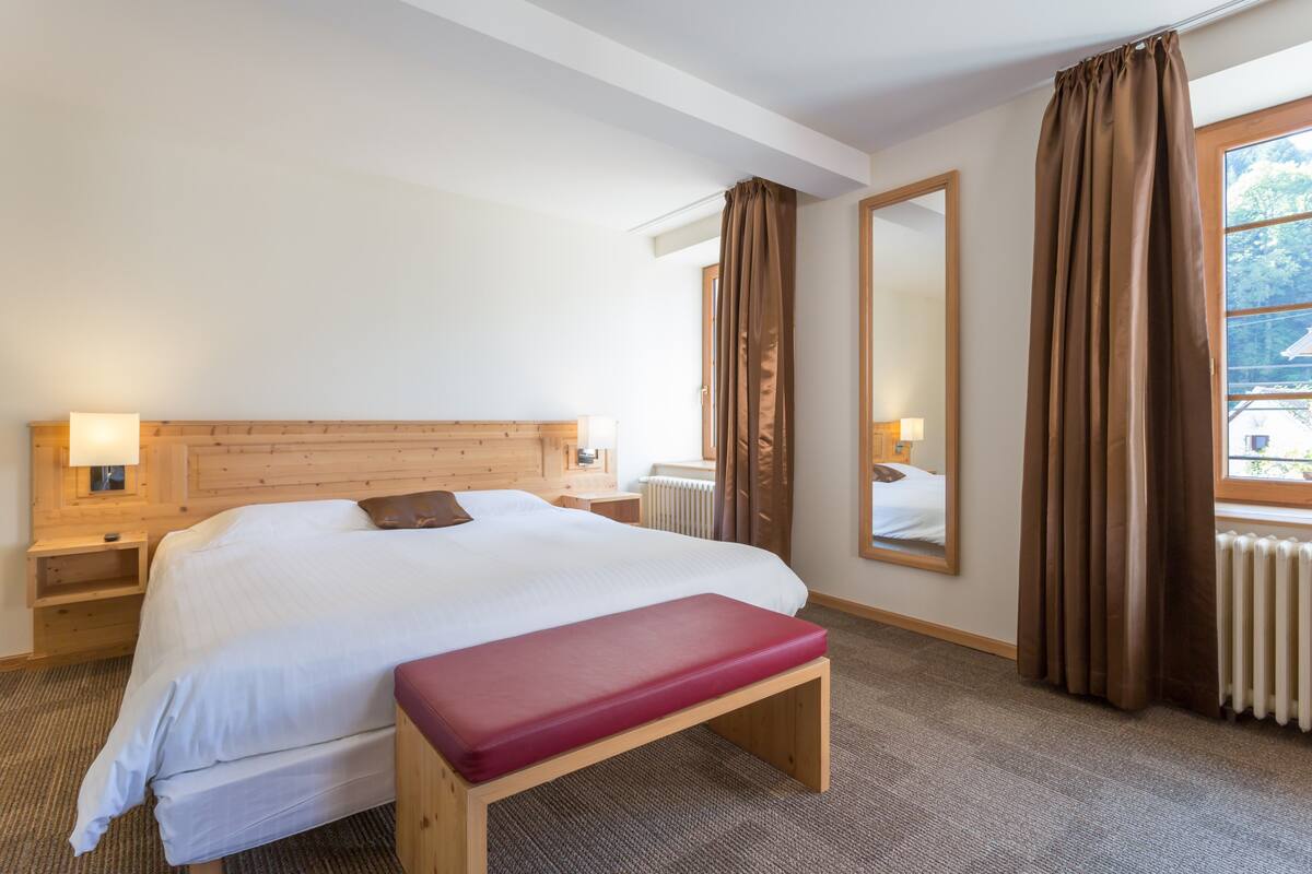 Habitación de hotel con una cama grande, cabecera de madera, un banco, mesita de noche, un espejo y una ventana con cortinas.