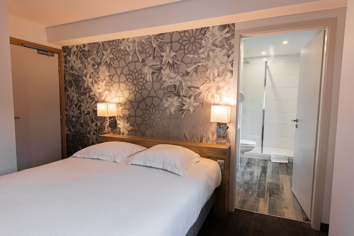 Habitación de hotel con cama, dos lámparas de noche, pared con estampado floral y zona de ducha visible a través de una puerta abierta.