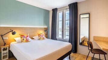 Ett hotellrum med en stor säng, ett skrivbord, en stol, en lampa och ett fönster med gardiner.