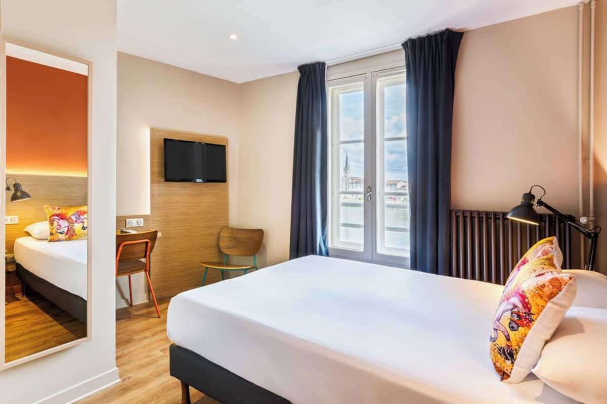 A hotel room with a bed, a desk, a chair, a TV, and a window with curtains.